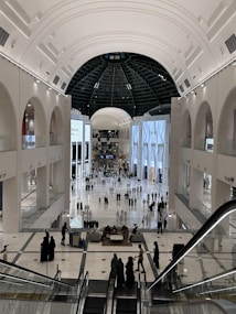 A spacious and modern indoor shopping mall featuring large, bright advertisements for luxury brands such as Prada and Dior. The mall has a high, arched ceiling with a central dome and multiple levels, connected by escalators. There are numerous people walking around, shopping, and socializing.