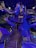A large, intricately detailed artificial tree structure indoors, with wooden stairs winding up to multiple levels and platforms. Blue and purple lighting creates a magical atmosphere, highlighting the textures and details of the tree's bark. The sign 'Tinkertree' is visible, indicating the theme or attraction name. The area beneath the tree is open, allowing for exploration and play.