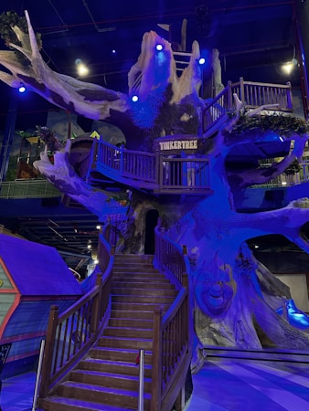 A large, intricately detailed artificial tree structure indoors, with wooden stairs winding up to multiple levels and platforms. Blue and purple lighting creates a magical atmosphere, highlighting the textures and details of the tree's bark. The sign 'Tinkertree' is visible, indicating the theme or attraction name. The area beneath the tree is open, allowing for exploration and play.
