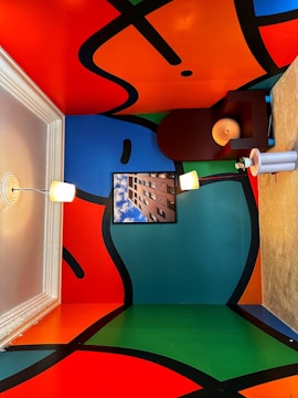 A vibrant interior space features a ceiling and walls painted with large abstract shapes in bright colors, framed by black lines. A white pendant light and a table lamp provide illumination. A framed photograph of a building against a blue sky is displayed on one of the walls. A small table holds a decorative object.