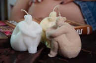 Three sculpted candles shaped like seated human figures are placed on a brown fabric surface. Two of the candles are white, while the third one has a beige tone. The background includes a glimpse of a person's hand resting on a book with a colorful cover and a denim fabric.