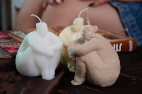 Three sculpted candles shaped like seated human figures are placed on a brown fabric surface. Two of the candles are white, while the third one has a beige tone. The background includes a glimpse of a person's hand resting on a book with a colorful cover and a denim fabric.