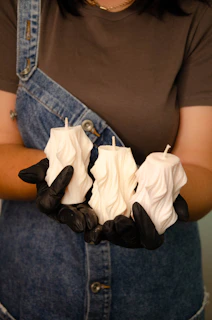 A person wearing a denim overall dress and gloves holds three sculpted white candles. The candles have a twisted, textured design.