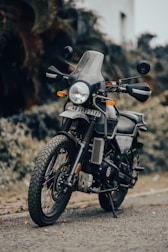 A black motorcycle is parked on the side of a road with a reflective front windshield and a visible license plate. The background is indistinct, featuring blurred vegetation.