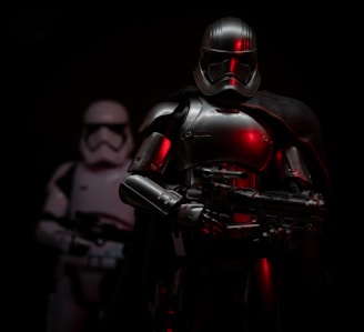 A figure in dark, metallic armor stands prominently with a reflective surface catching dim light and hints of red. Another similar figure appears slightly in the background, blurred. The setting is dark, creating a mysterious and ominous atmosphere.