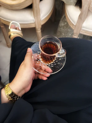 A hand wearing hanjiara rings holding a steaming cup of tea by a window.