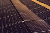 A series of solar panels arranged in rows, capturing sunlight. The panels have a grid-like pattern and are placed on a dark, gravel-covered surface. The symmetry and alignment of the panels give a sense of order and efficiency.