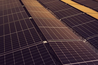 A series of solar panels arranged in rows, capturing sunlight. The panels have a grid-like pattern and are placed on a dark, gravel-covered surface. The symmetry and alignment of the panels give a sense of order and efficiency.