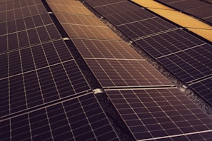 A series of solar panels arranged in rows, capturing sunlight. The panels have a grid-like pattern and are placed on a dark, gravel-covered surface. The symmetry and alignment of the panels give a sense of order and efficiency.
