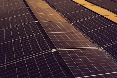 A series of solar panels arranged in rows, capturing sunlight. The panels have a grid-like pattern and are placed on a dark, gravel-covered surface. The symmetry and alignment of the panels give a sense of order and efficiency.