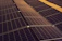 A series of solar panels arranged in rows, capturing sunlight. The panels have a grid-like pattern and are placed on a dark, gravel-covered surface. The symmetry and alignment of the panels give a sense of order and efficiency.