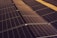 A series of solar panels arranged in rows, capturing sunlight. The panels have a grid-like pattern and are placed on a dark, gravel-covered surface. The symmetry and alignment of the panels give a sense of order and efficiency.