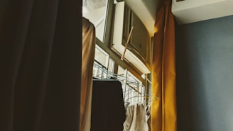 An indoor scene featuring an air conditioning unit installed above a window, with curtains partially drawn to the side. A clothes rack with hangers is visible, positioned near the window. The lighting suggests a soft, possibly morning or late afternoon ambiance.