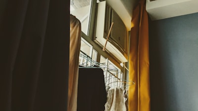 An indoor scene featuring an air conditioning unit installed above a window, with curtains partially drawn to the side. A clothes rack with hangers is visible, positioned near the window. The lighting suggests a soft, possibly morning or late afternoon ambiance.