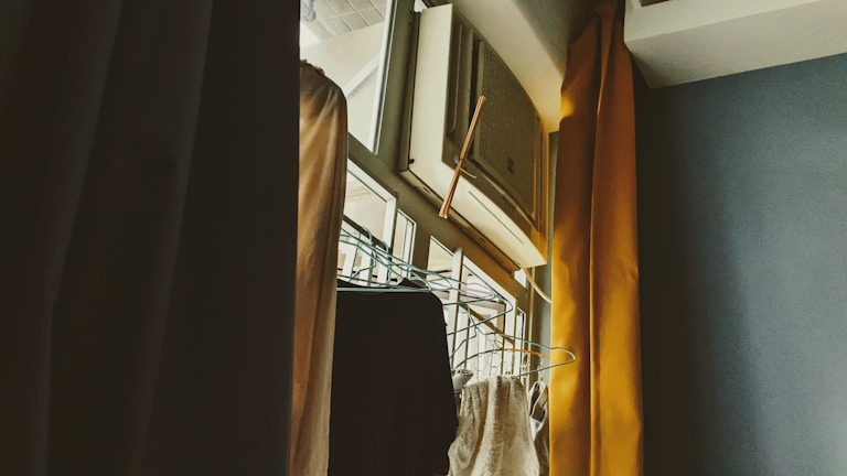 An indoor scene featuring an air conditioning unit installed above a window, with curtains partially drawn to the side. A clothes rack with hangers is visible, positioned near the window. The lighting suggests a soft, possibly morning or late afternoon ambiance.
