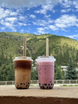 Two cups of bubble tea with lids and straws placed on a ledge, set against a backdrop of lush green hills and a partly cloudy blue sky.