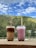 Two cups of bubble tea with lids and straws placed on a ledge, set against a backdrop of lush green hills and a partly cloudy blue sky.