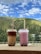 Two cups of bubble tea with lids and straws placed on a ledge, set against a backdrop of lush green hills and a partly cloudy blue sky.