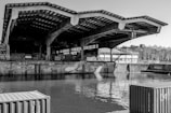 A large, industrial structure with a distinctive, angular roof extends over a waterway. The building appears to be a warehouse or dock, surrounded by walls with graffiti. Shipping containers are visible in the foreground, and the water reflects the structure. In the background, leafless trees and industrial buildings line the horizon.