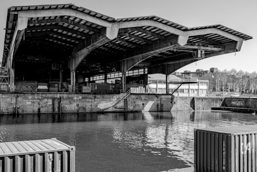 A large, industrial structure with a distinctive, angular roof extends over a waterway. The building appears to be a warehouse or dock, surrounded by walls with graffiti. Shipping containers are visible in the foreground, and the water reflects the structure. In the background, leafless trees and industrial buildings line the horizon.