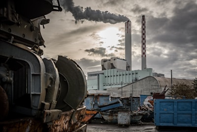 A large industrial facility with tall chimneys emitting thick smoke against a cloudy sky. Old machinery and various industrial containers are scattered in the foreground.