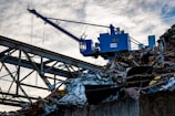 A blue industrial crane is positioned atop a large pile of scrap metal. The structure is silhouetted against a cloudy sky, with metal beams and wires forming part of the complex environment.