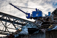 A blue industrial crane is positioned atop a large pile of scrap metal. The structure is silhouetted against a cloudy sky, with metal beams and wires forming part of the complex environment.