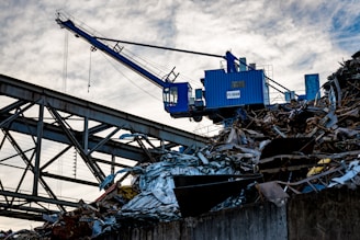 A blue industrial crane is positioned atop a large pile of scrap metal. The structure is silhouetted against a cloudy sky, with metal beams and wires forming part of the complex environment.