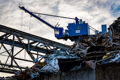 A blue industrial crane is positioned atop a large pile of scrap metal. The structure is silhouetted against a cloudy sky, with metal beams and wires forming part of the complex environment.