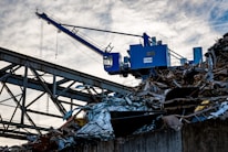 A blue industrial crane is positioned atop a large pile of scrap metal. The structure is silhouetted against a cloudy sky, with metal beams and wires forming part of the complex environment.