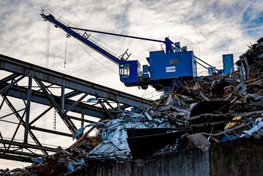 A blue industrial crane is positioned atop a large pile of scrap metal. The structure is silhouetted against a cloudy sky, with metal beams and wires forming part of the complex environment.