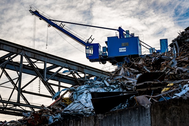 A blue industrial crane is positioned atop a large pile of scrap metal. The structure is silhouetted against a cloudy sky, with metal beams and wires forming part of the complex environment.