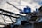 A blue industrial crane is positioned atop a large pile of scrap metal. The structure is silhouetted against a cloudy sky, with metal beams and wires forming part of the complex environment.