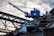 A blue industrial crane is positioned atop a large pile of scrap metal. The structure is silhouetted against a cloudy sky, with metal beams and wires forming part of the complex environment.