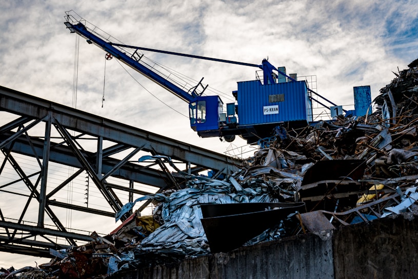 A blue industrial crane is positioned atop a large pile of scrap metal. The structure is silhouetted against a cloudy sky, with metal beams and wires forming part of the complex environment.