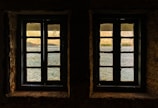Two windows with muntin bars are set in a stone wall, offering a view of a serene body of water with reflections. Beyond the water, a distant shoreline with trees and buildings is visible under a gentle, warm sky.