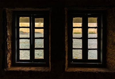 Two windows with muntin bars are set in a stone wall, offering a view of a serene body of water with reflections. Beyond the water, a distant shoreline with trees and buildings is visible under a gentle, warm sky.