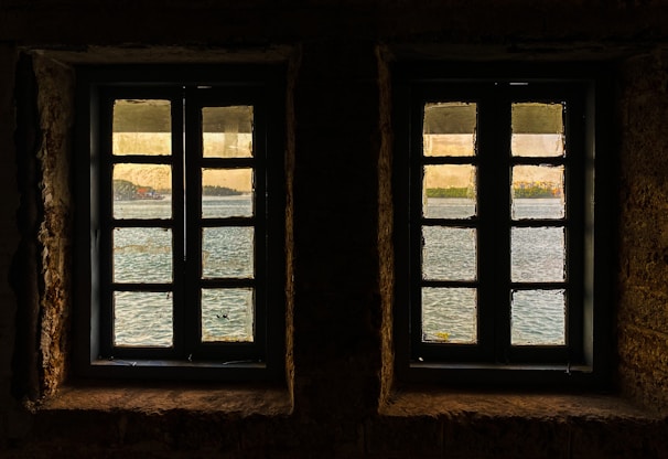 Two windows with muntin bars are set in a stone wall, offering a view of a serene body of water with reflections. Beyond the water, a distant shoreline with trees and buildings is visible under a gentle, warm sky.