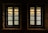 Two windows with muntin bars are set in a stone wall, offering a view of a serene body of water with reflections. Beyond the water, a distant shoreline with trees and buildings is visible under a gentle, warm sky.