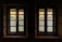 Two windows with muntin bars are set in a stone wall, offering a view of a serene body of water with reflections. Beyond the water, a distant shoreline with trees and buildings is visible under a gentle, warm sky.