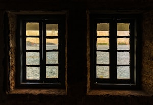 Two windows with muntin bars are set in a stone wall, offering a view of a serene body of water with reflections. Beyond the water, a distant shoreline with trees and buildings is visible under a gentle, warm sky.