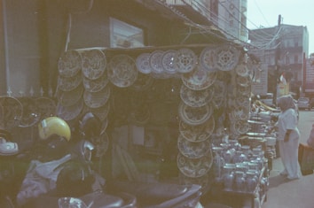 A market stall displaying a collection of decorative plates with intricate patterns hanging on the sides. Below the plates, there are shelves filled with glass jars and other items. A person wearing a headscarf and white attire is browsing the items. Motorcycles and a yellow helmet are visible in the foreground, and the surroundings suggest an urban market setting.