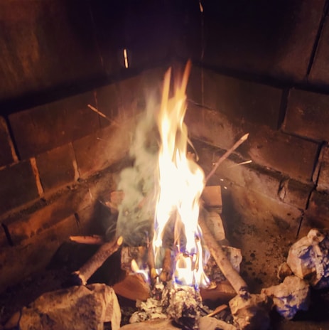 An indoor fireplace with burning wood creates a warm glow, casting light on surrounding bricks. Smoke rises gently from the flames.