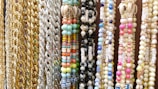 A variety of colorful beaded and chain necklaces are hanging vertically. The collection features gold and silver chains alongside beaded designs in multiple colors including pastel blues, pinks, greens, and yellows. Some beads have letters spelling out words like 'HAPPY'. There are also shells embedded in some of the necklaces.