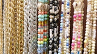 A variety of colorful beaded and chain necklaces are hanging vertically. The collection features gold and silver chains alongside beaded designs in multiple colors including pastel blues, pinks, greens, and yellows. Some beads have letters spelling out words like 'HAPPY'. There are also shells embedded in some of the necklaces.