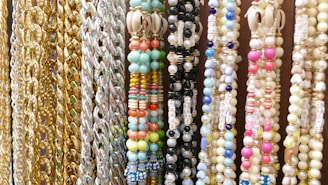 A variety of colorful beaded and chain necklaces are hanging vertically. The collection features gold and silver chains alongside beaded designs in multiple colors including pastel blues, pinks, greens, and yellows. Some beads have letters spelling out words like 'HAPPY'. There are also shells embedded in some of the necklaces.