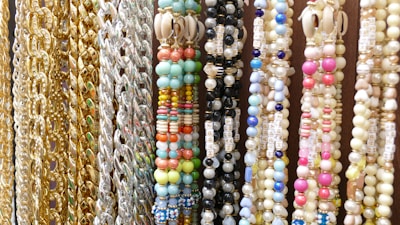 A variety of colorful beaded and chain necklaces are hanging vertically. The collection features gold and silver chains alongside beaded designs in multiple colors including pastel blues, pinks, greens, and yellows. Some beads have letters spelling out words like 'HAPPY'. There are also shells embedded in some of the necklaces.