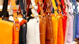 A row of colorful leather handbags displayed in a variety of vibrant shades ranging from orange, black, white, and brown to yellow, red, purple, and pink. The bags are neatly hung by their straps, highlighting their design and metallic buckles.