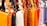 A row of colorful leather handbags displayed in a variety of vibrant shades ranging from orange, black, white, and brown to yellow, red, purple, and pink. The bags are neatly hung by their straps, highlighting their design and metallic buckles.