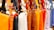 A row of colorful leather handbags displayed in a variety of vibrant shades ranging from orange, black, white, and brown to yellow, red, purple, and pink. The bags are neatly hung by their straps, highlighting their design and metallic buckles.
